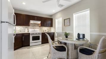 A kitchen with dark wooden cabinets, a white refrigerator, and a built-in oven. There is a dining table with wicker chairs and a potted plant.