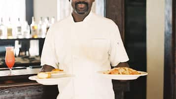 Een kok in een witte uniform met borden eten en een drankje, staand in een barruimte met betegelde spatwand en houten details.