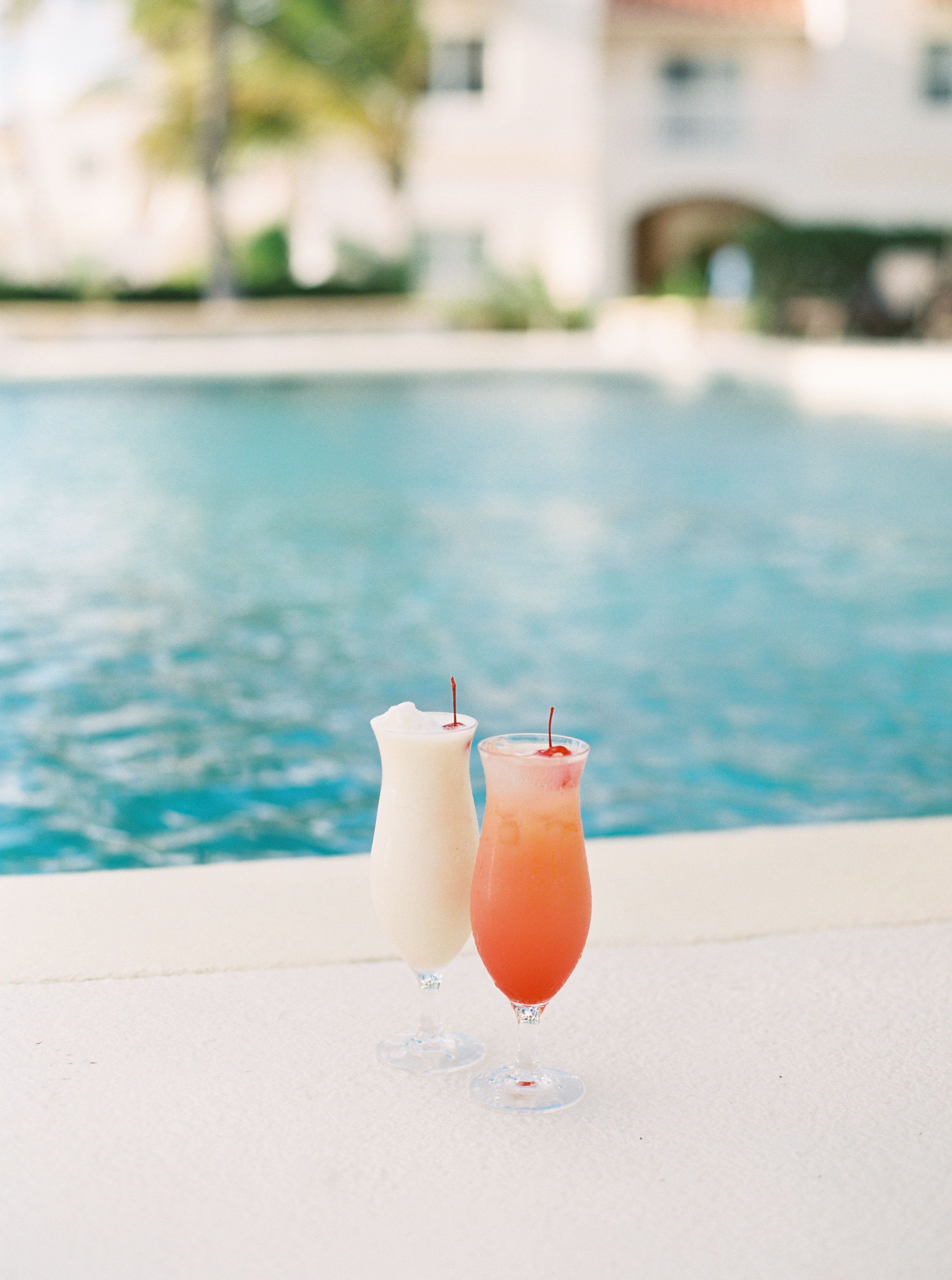 Two drinks on a poolside table with a pool and a building in the background.