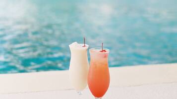 Two drinks on a poolside table with a pool and a building in the background.