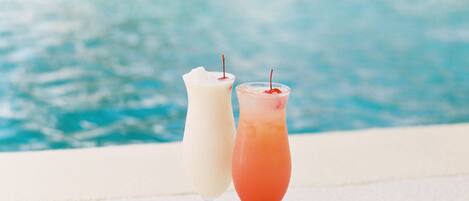 Two drinks on a poolside table with a pool and a building in the background.