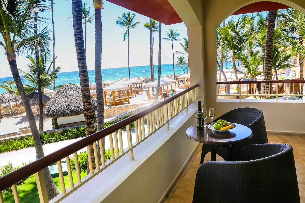 A balcony with a view of the beach, palm trees, and a table with a fruit bowl and glasses.