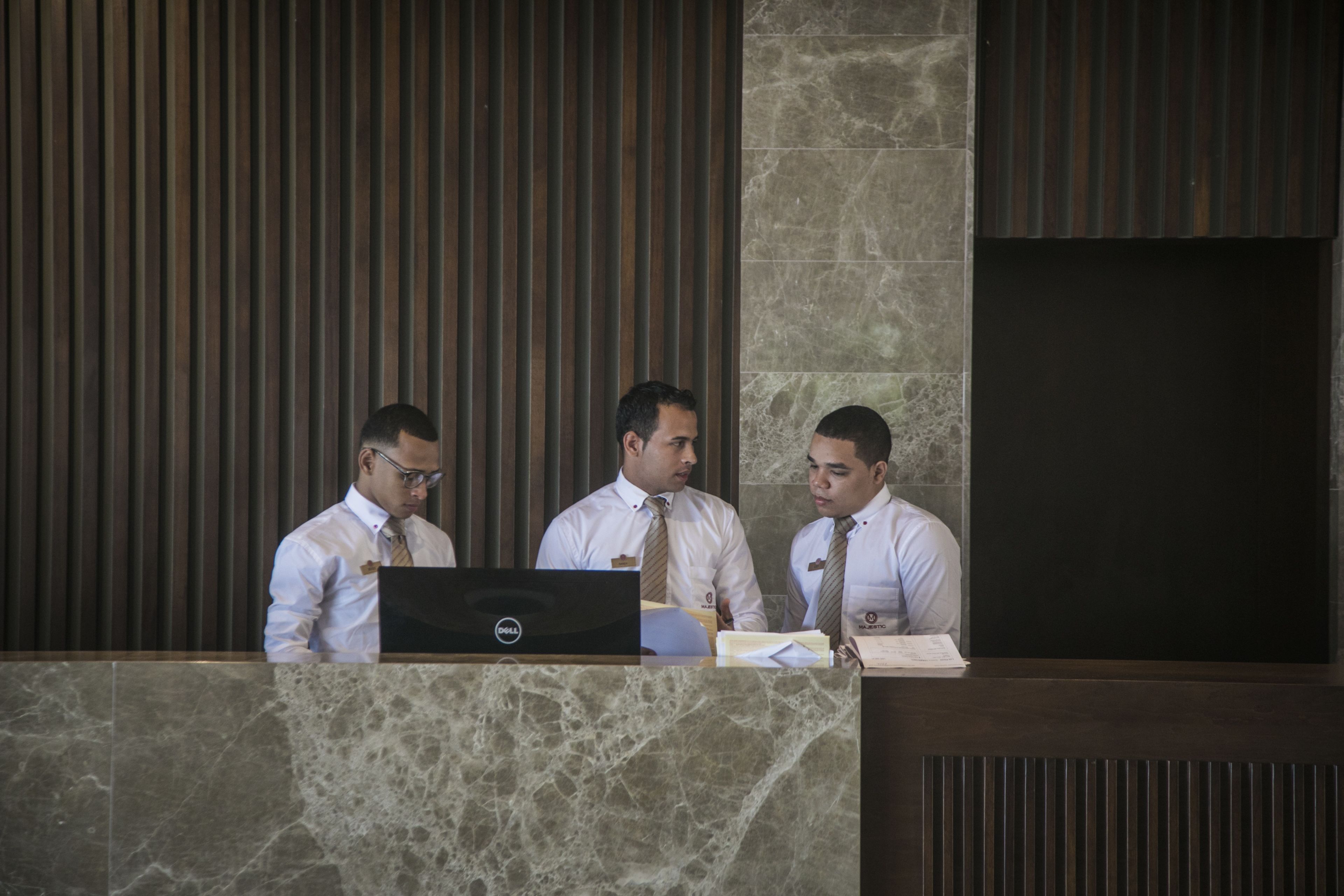 Trois hommes sont assis derrière un comptoir d’accueil, devant un ordinateur portable, des documents et un bloc-notes.