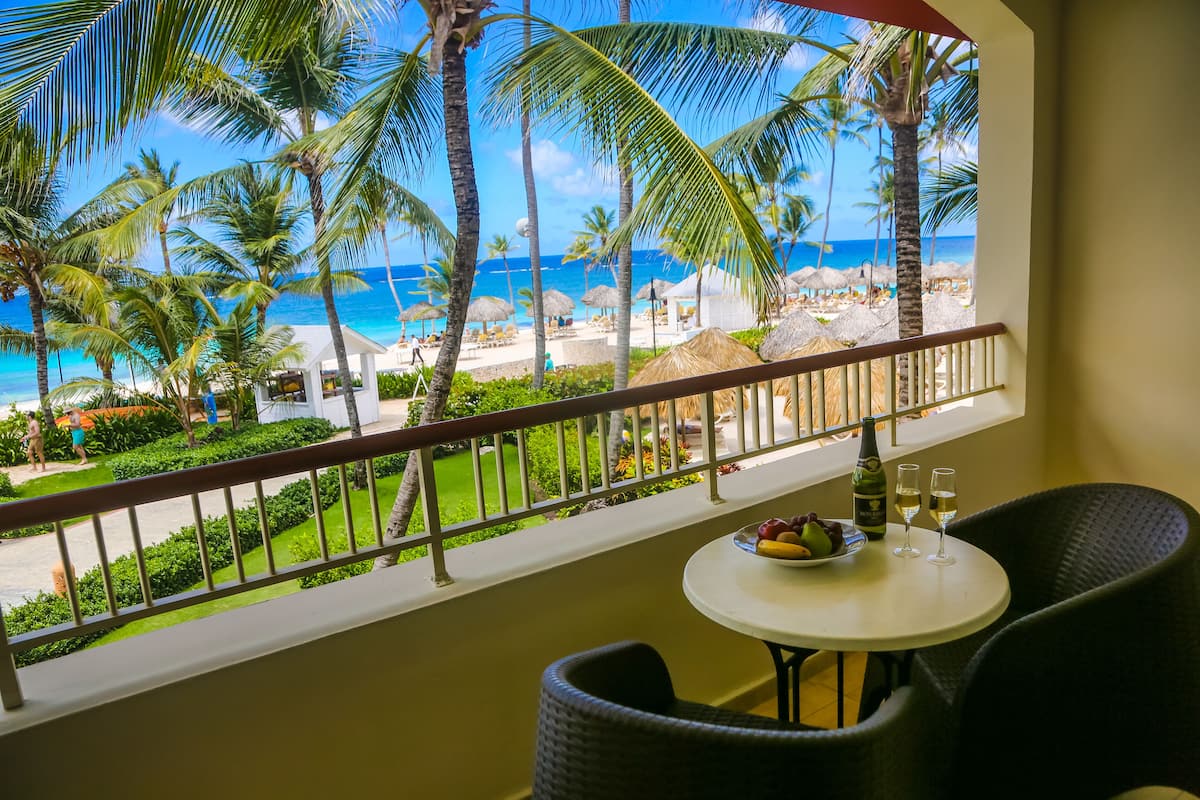 A balcony with a view of a beach, a table with a fruit bowl and glasses, and a bottle of wine.