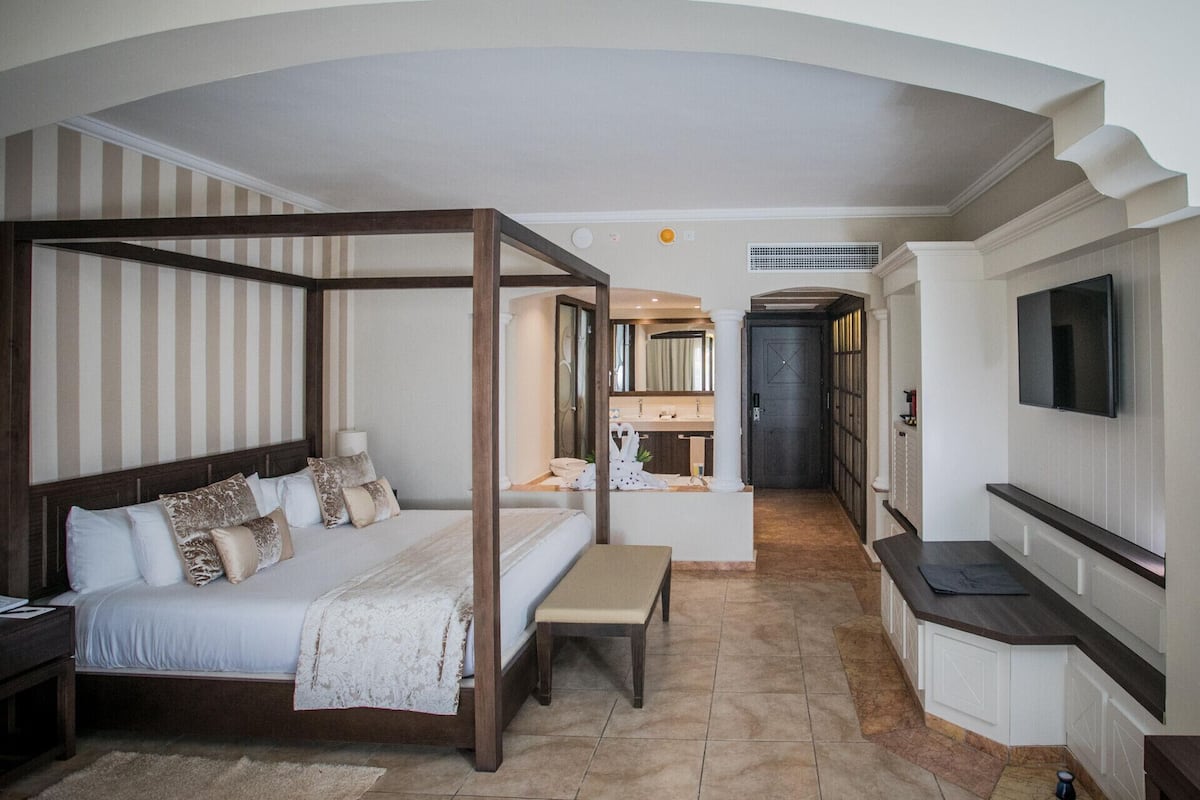 A four-poster bed with a bench, a bathroom with a mirror and sink, and a TV mounted on the wall.