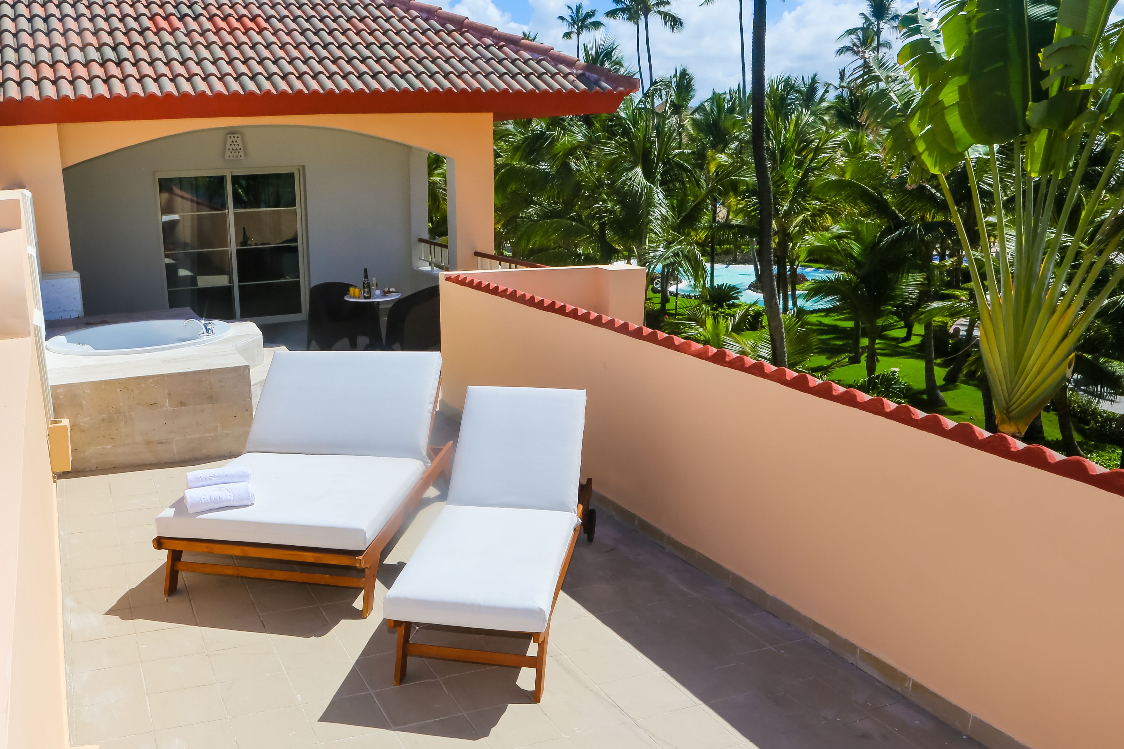 Un balcón con dos sillones blancos, un jacuzzi y vistas a una piscina y palmeras.