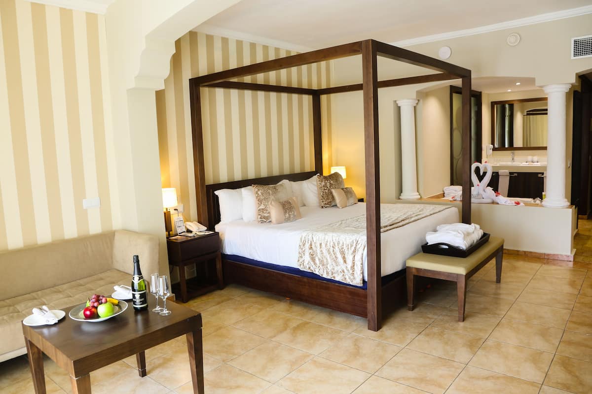A four-poster bed with a wooden frame, a bedside table with a bottle and glasses, a bench with towels, a fruit bowl, and a nightstand with a lamp.