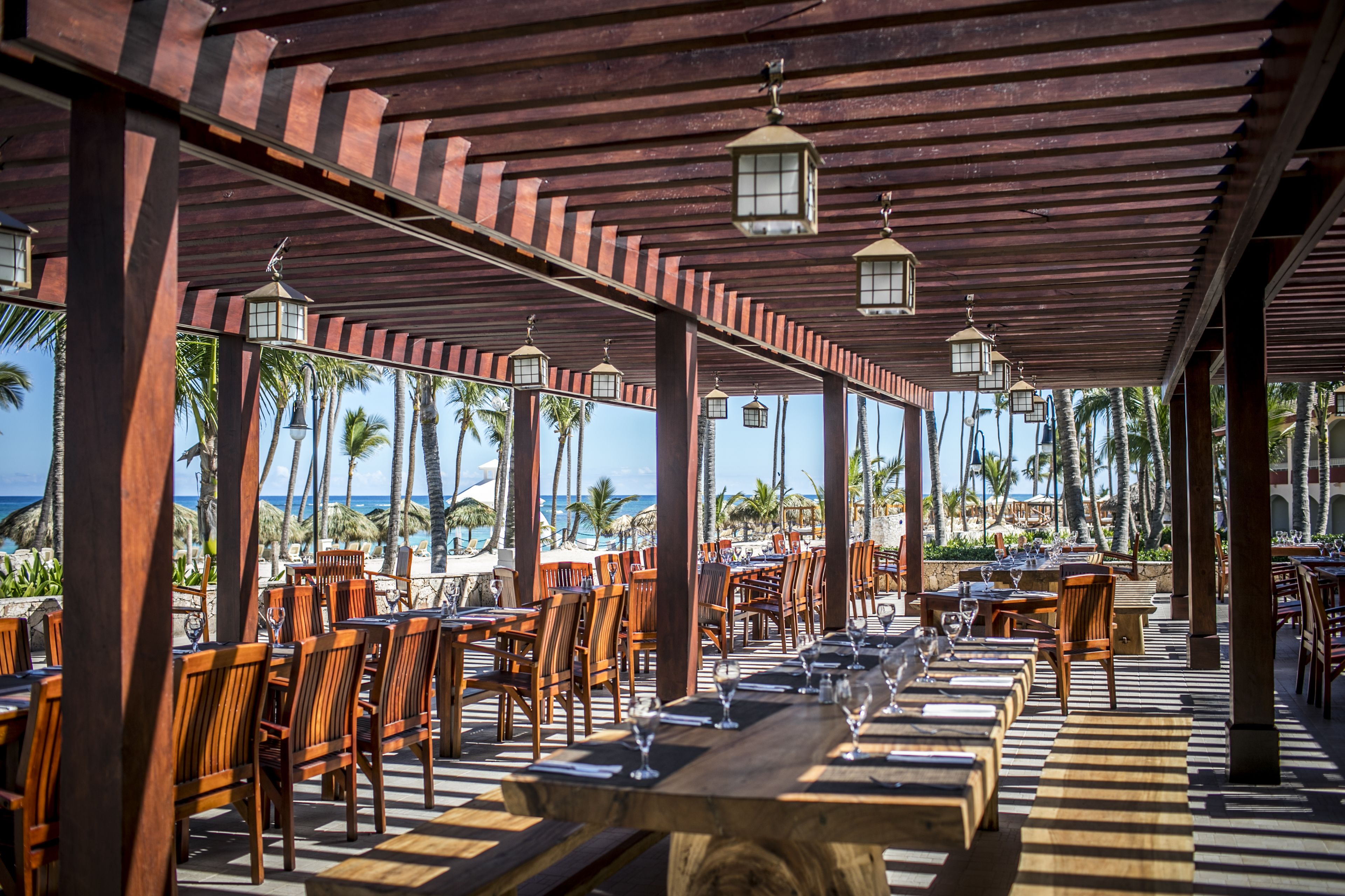 An outdoor dining area with wooden tables and chairs, surrounded by palm trees and a view of the ocean.