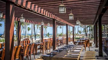 An outdoor dining area with wooden tables and chairs, surrounded by palm trees and a view of the ocean.
