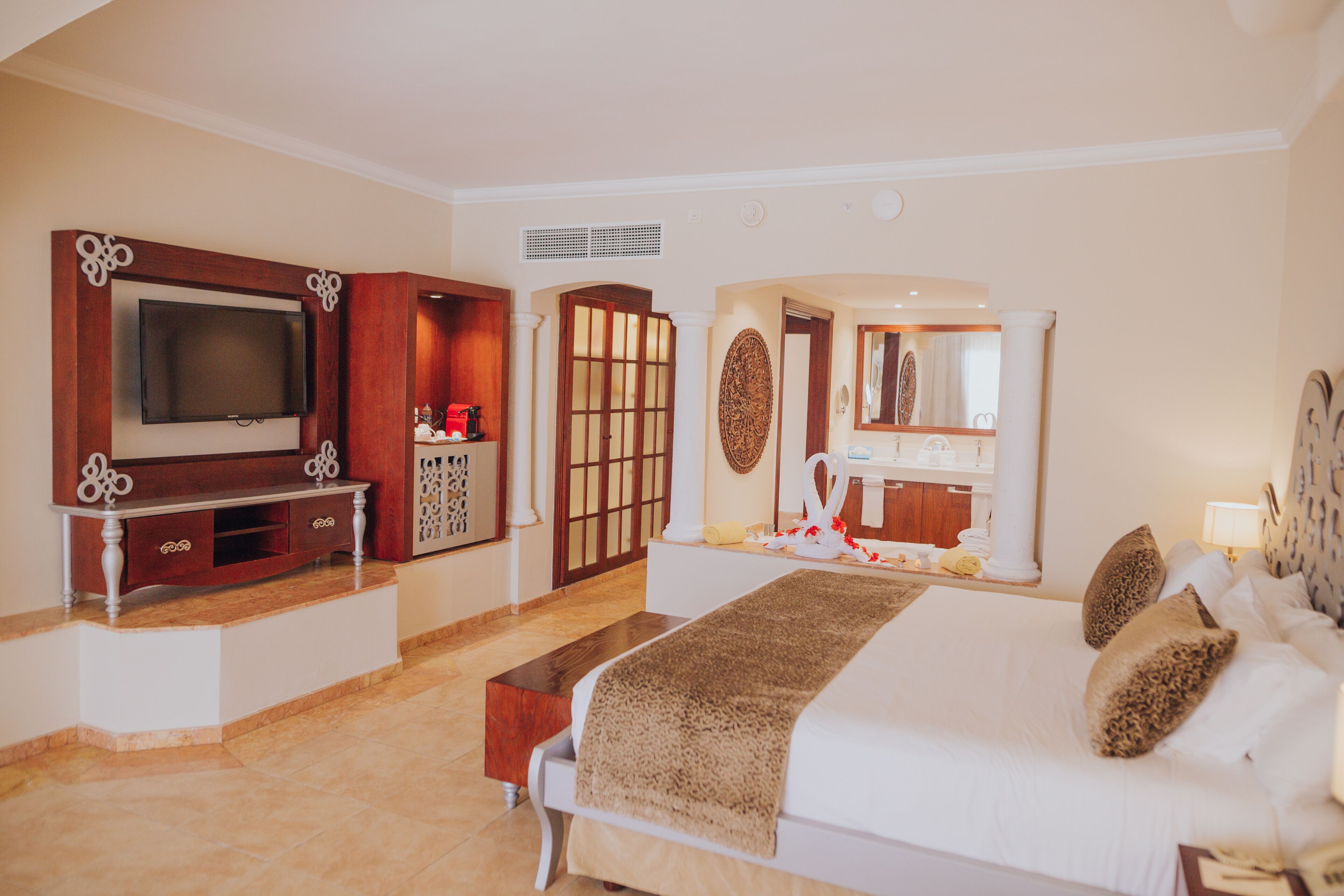 Habitación de hotel con una cama grande, un televisor montado en un mueble de madera, un tocador con espejo y un pequeño rincón de descanso.