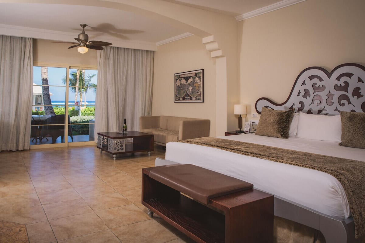 A hotel room with a bed, a desk, a chair, a coffee table, a ceiling fan, and a view of the ocean.