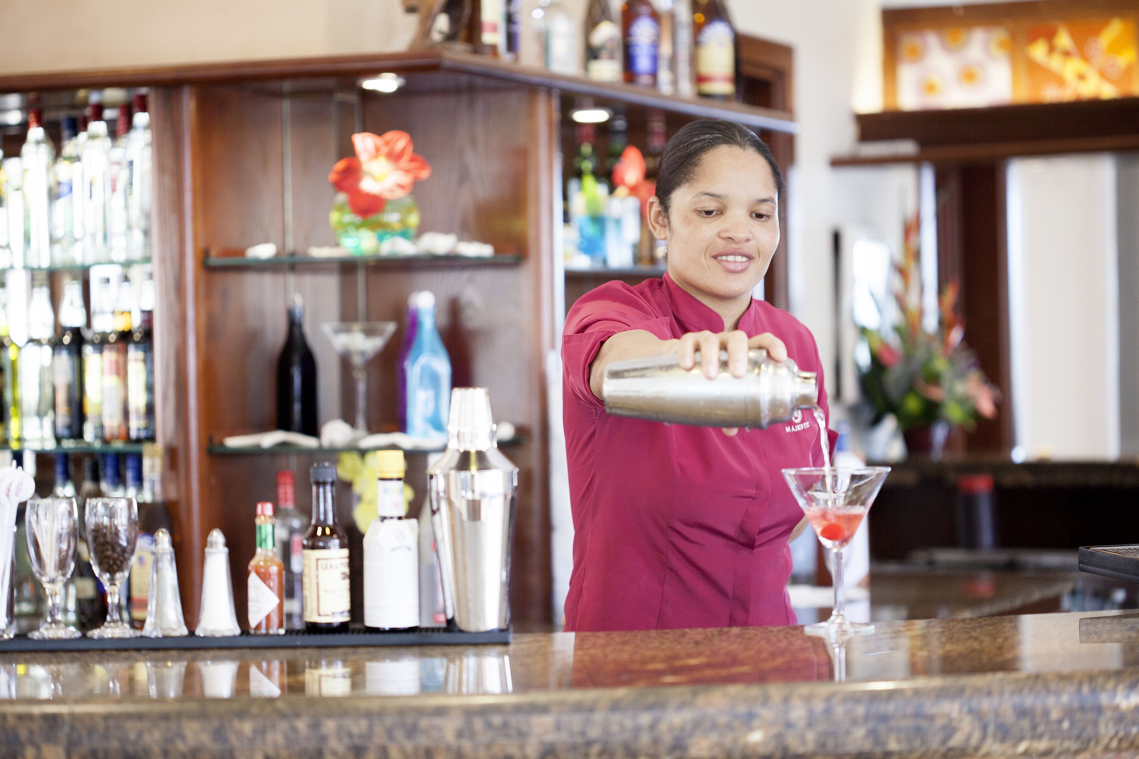 A bartender preparing a drink at a bar with shelves of bottles and glasses in the background.
