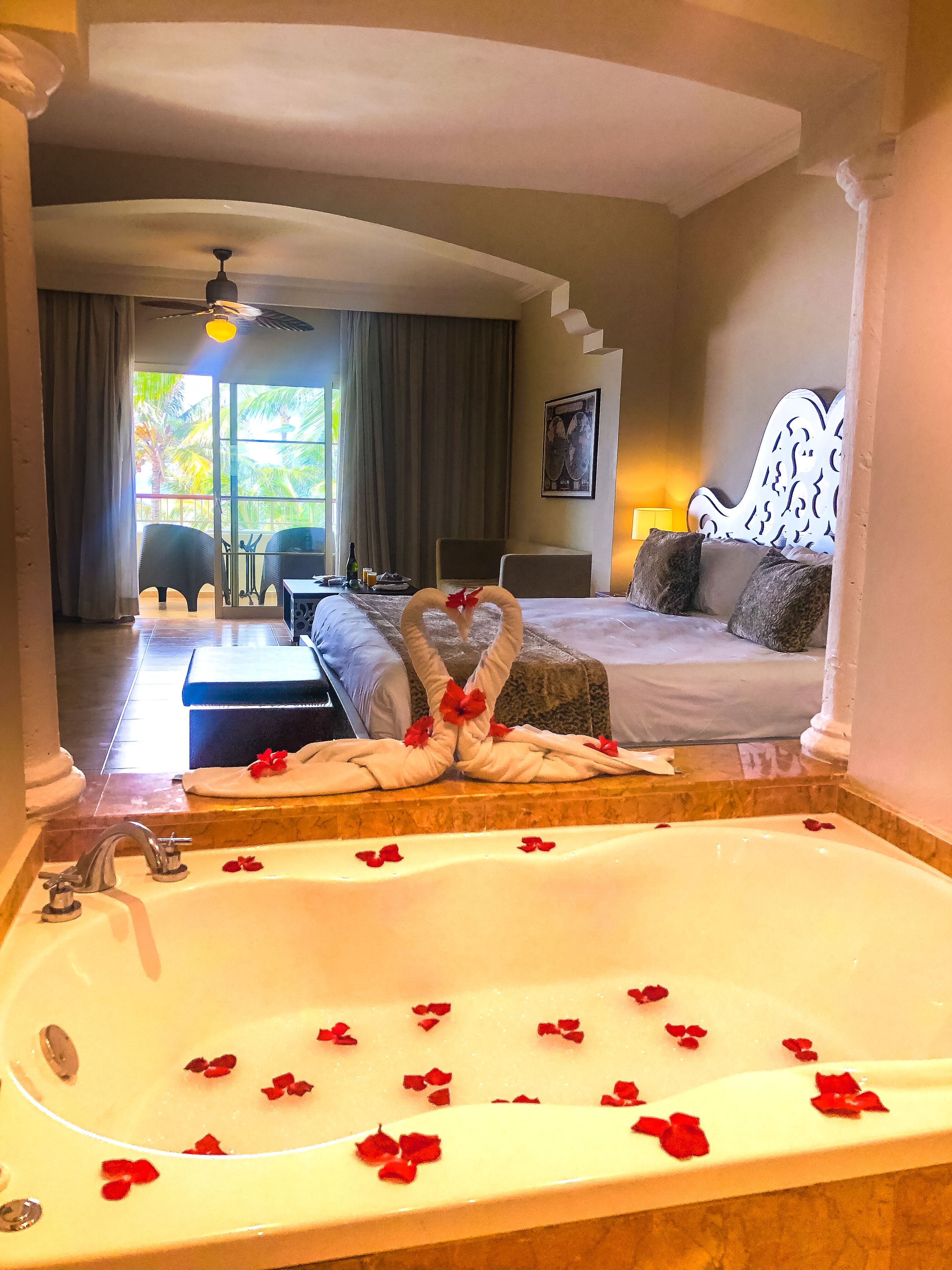 Een hotelkamer met een groot bad gevuld met rozenblaadjes, een bed met een wit hoofdeinde, en een balkon met stoelen en een tafel.