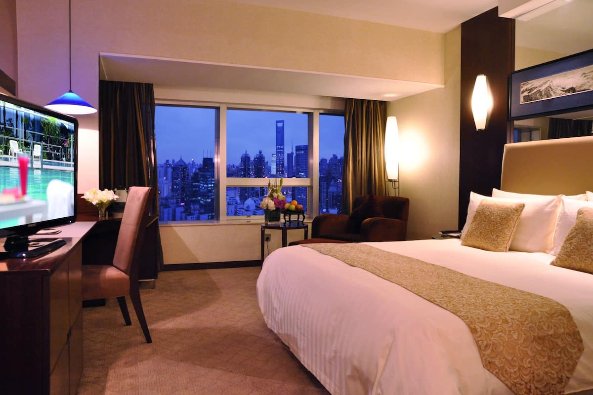 Habitación de hotel con una cama grande, un escritorio con computadora, una silla, una mesita con un florero y vistas a la silueta urbana a través de amplias ventanas.