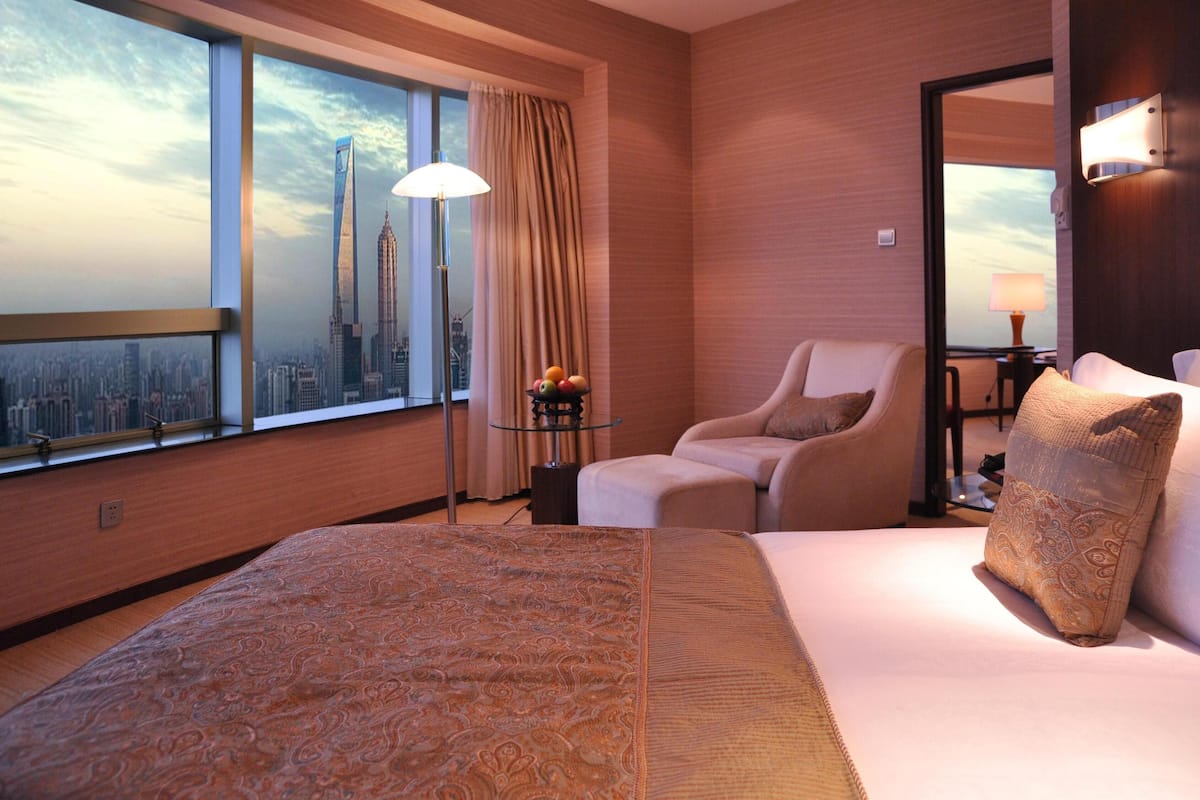 Habitación de hotel con una cama grande, una silla, una mesita con lámpara y vistas a la ciudad a través de una ventana.