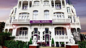 A large, white building with a sign reading "HOTEL COREI." It features multiple balconies, large windows, and a central entrance with a decorative archway. - Hotel Corel (The Hague)