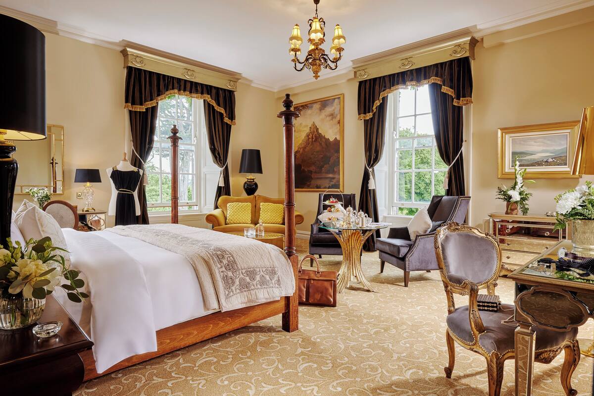 A spacious bedroom with a large bed, a sitting area with a table and chairs, and a view of the outdoors through large windows.
