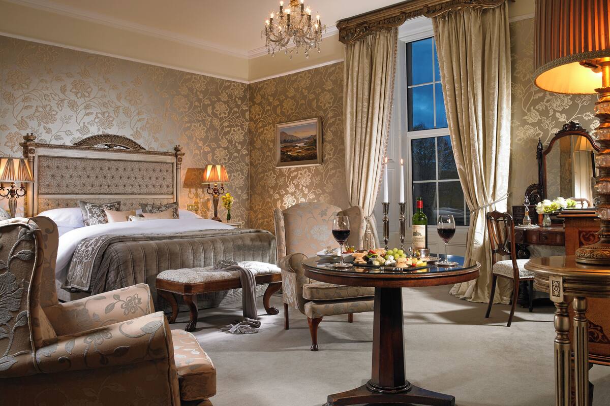 A luxurious bedroom with a large bed, ornate furniture, a chandelier, and a dining table set with wine glasses and a fruit platter.