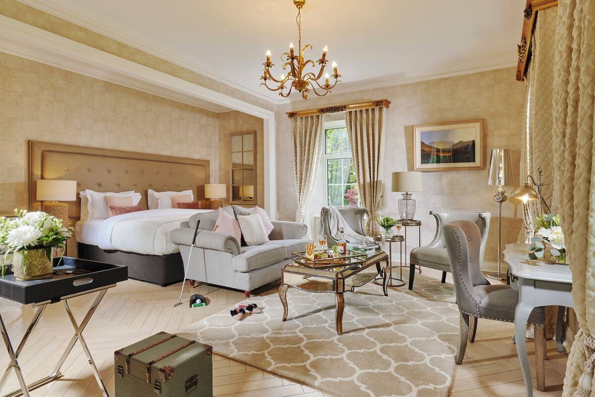 A hotel room with a large bed, a sofa, a coffee table, and a chair. There is a chandelier, a painting on the wall, and a window with curtains.