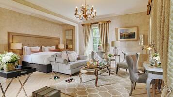 Habitación de hotel con una cama grande, un sofá, una mesa de centro y una silla. Se observa un candelabro, un cuadro en la pared y una ventana con cortinas.