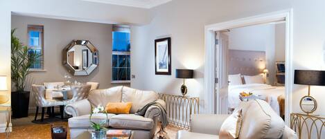 A spacious living room with a large sofa, a coffee table with books, a dining area with a table and chairs, and a bedroom visible through an open door.