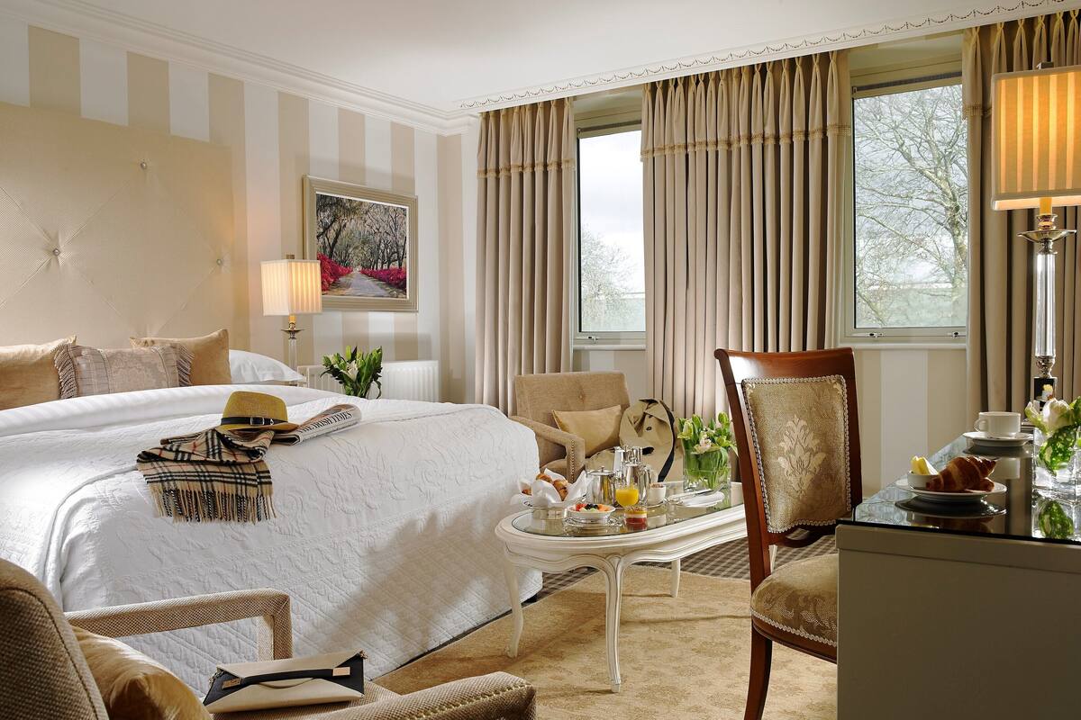 A hotel room with a large bed, a dining table with breakfast items, a chair, and a view of trees through the window.
