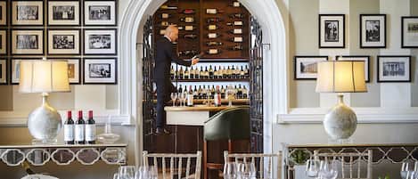 A bar area with a wine rack, a man standing by the bar, and a table set for dining with glasses and a vase of flowers.