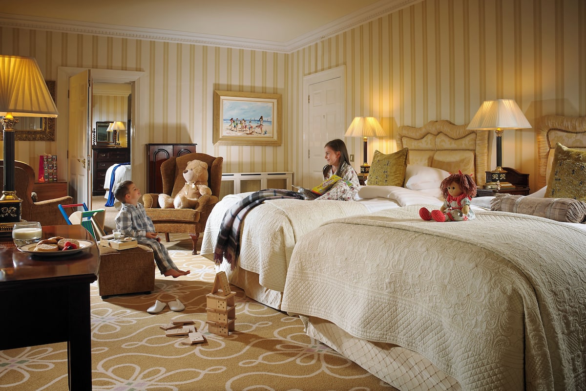 A hotel room with a large bed, a sitting area with a teddy bear, a dining table with breakfast items, and a painting on the wall.