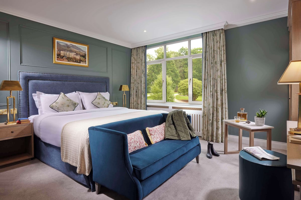 A bedroom with a large bed, a blue sofa, a nightstand, a table with a lamp, and a window with curtains.