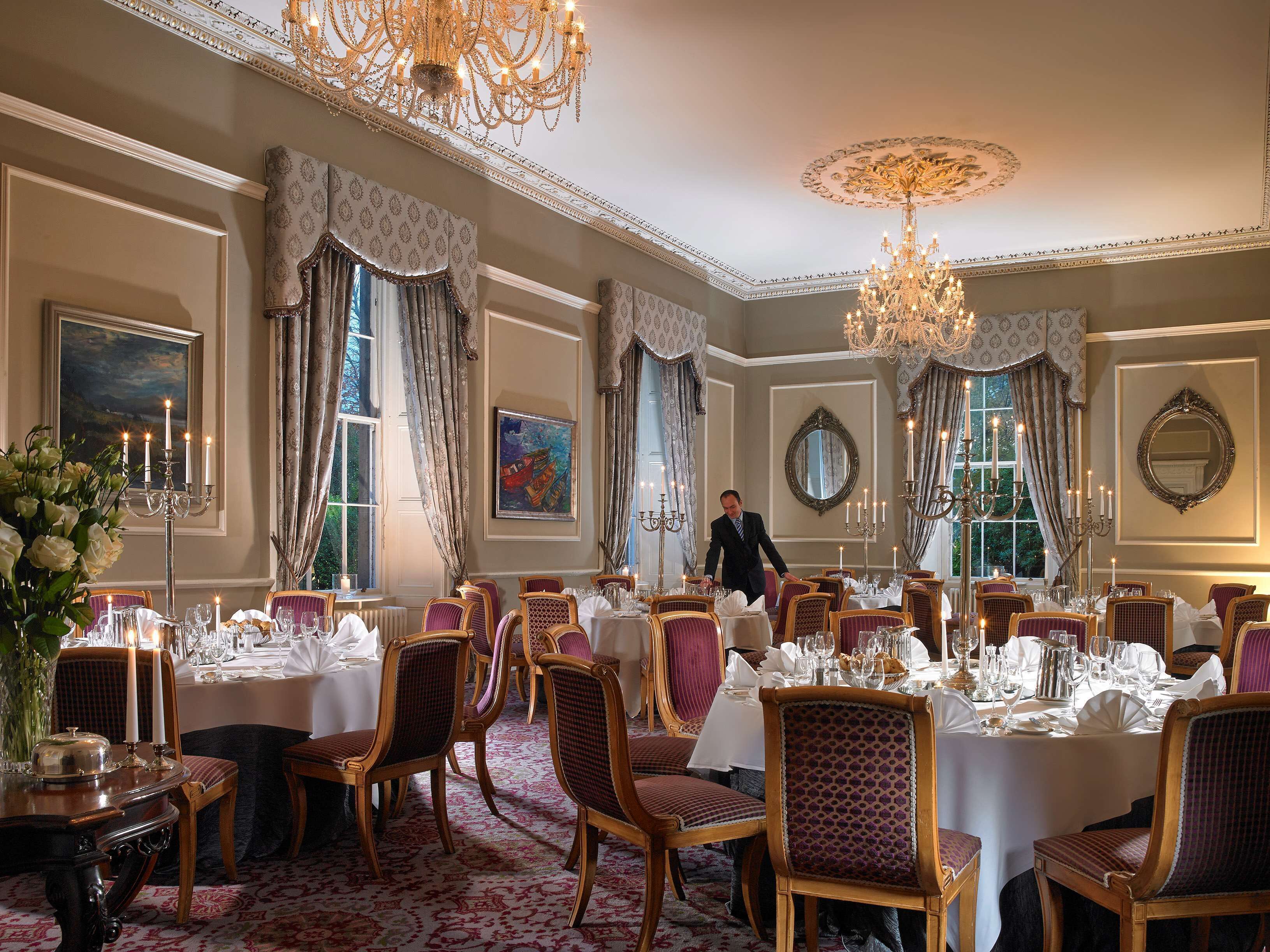 A formal dining room with multiple tables set for service, elegant chandeliers, and classic decor.