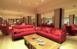 Un rebedor d'hotel espaiós amb un sofà seccional vermell gran, una taula de centre de fusta amb llibres i una zona de recepció al fons.