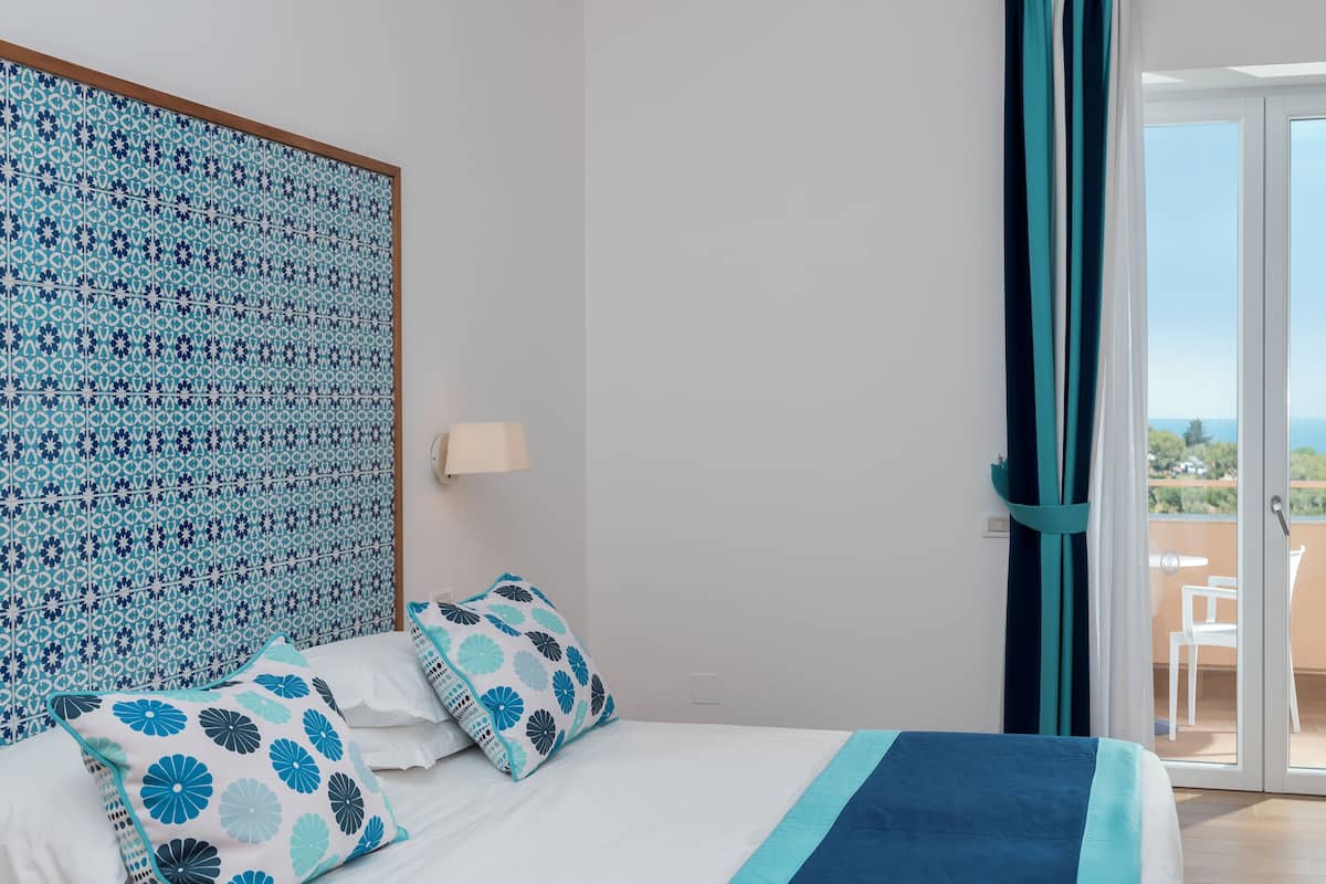 A bedroom with a patterned headboard, a bed with floral pillows, a nightstand, and a view of the outdoors through a sliding glass door.