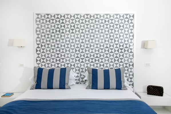 A modern hotel room with a geometric patterned headboard, a neatly made bed with blue and gray striped pillows, a white bedside table with a lamp, and a small Louis Vuitton bag on the nightstand. - Hotel Miramare (Sorrento)