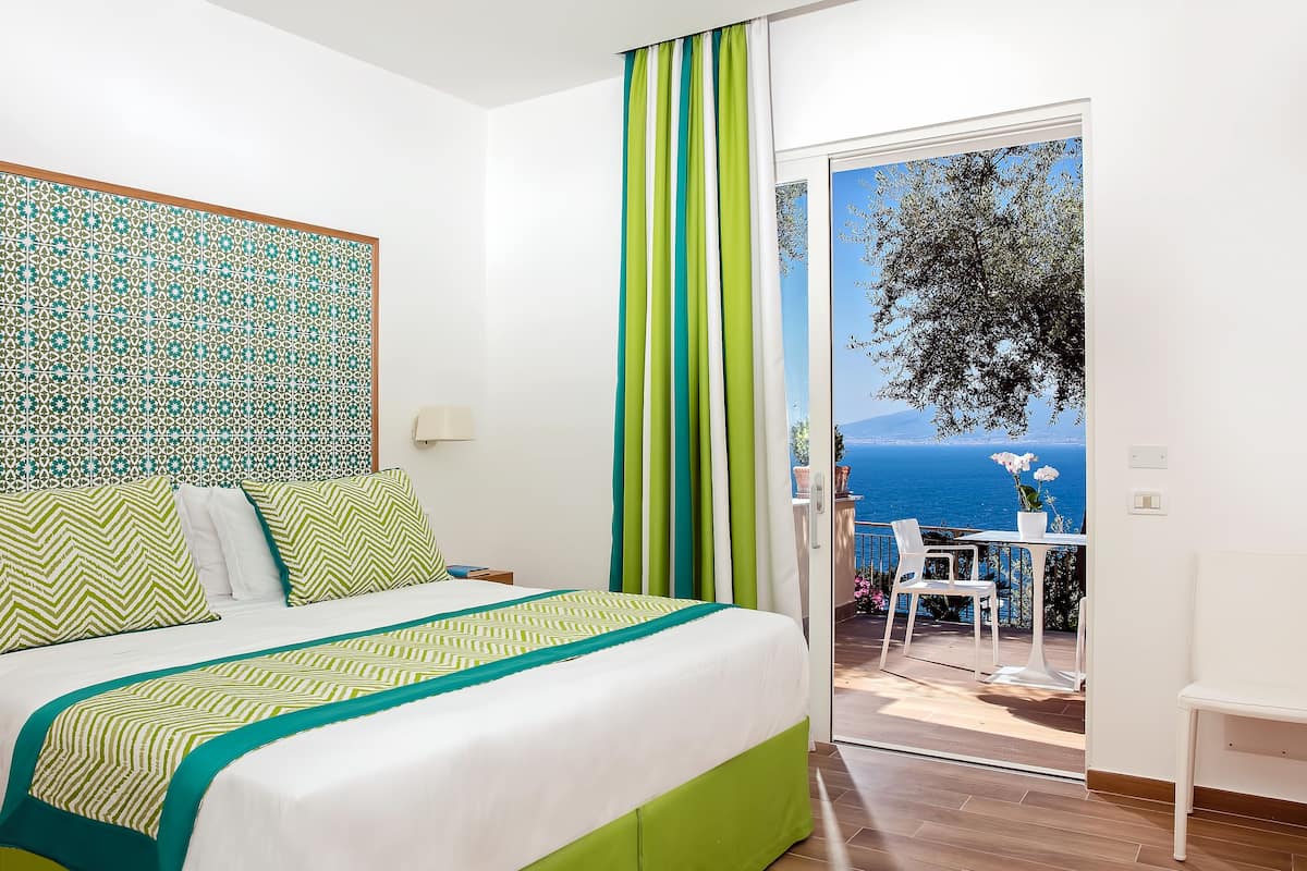 A hotel room with a bed, a view of the sea, and a balcony with outdoor seating.