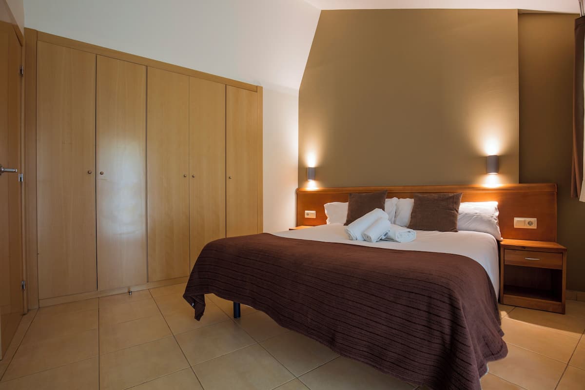 A bedroom with a large wardrobe, a bed with a brown bedspread, two bedside lamps, and a nightstand.