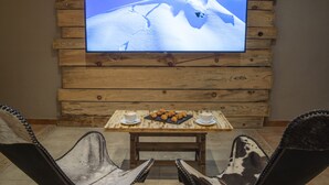 Pared de madera con un televisor de pantalla plana mostrando un paisaje nevado de montaña. Frente a ella, una mesa de madera con dos tazas y un plato de pasteles.