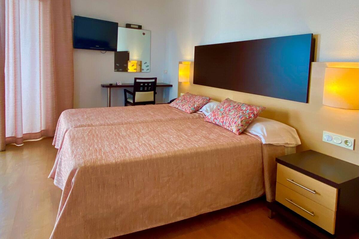 A hotel room with a double bed, bedside tables, a desk, a chair, a mirror, and a TV.