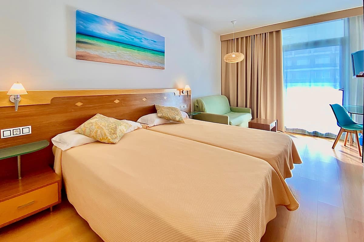 A hotel room with two beds, a green armchair, a TV, and a large window.