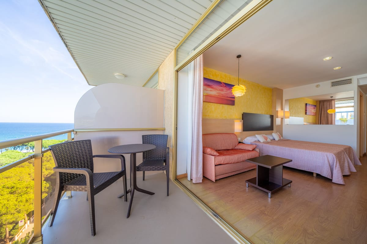 A balcony with wicker chairs and a small table, a bedroom with a bed, a TV, and a painting on the wall.