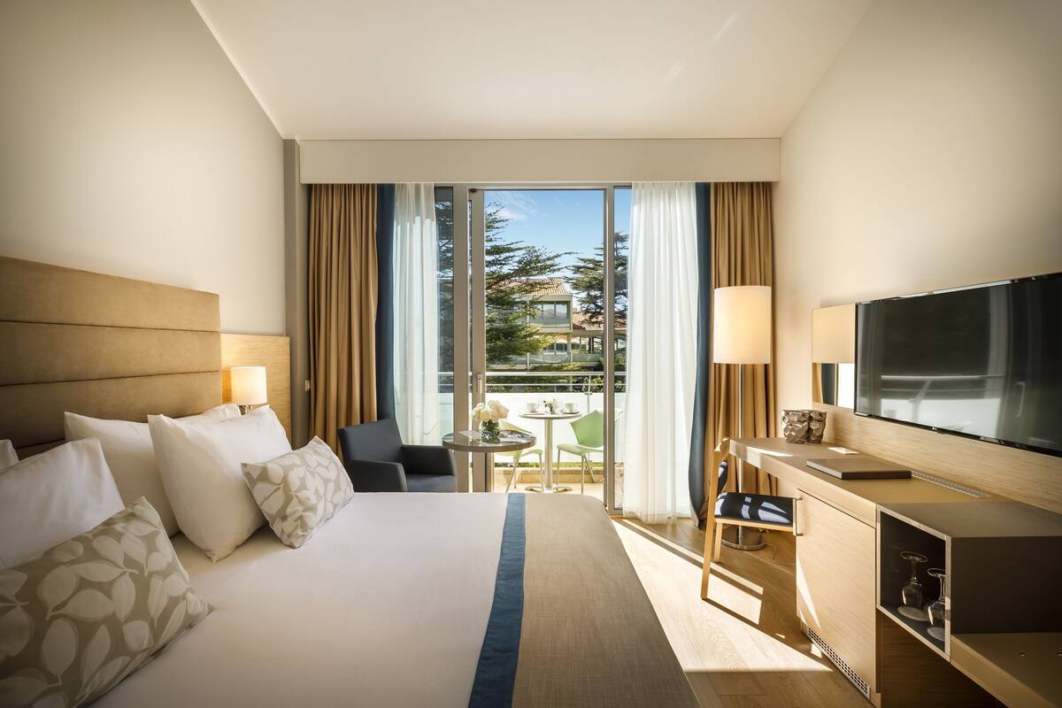 A hotel room with a large bed, a TV, a desk, and a balcony with a view of buildings.