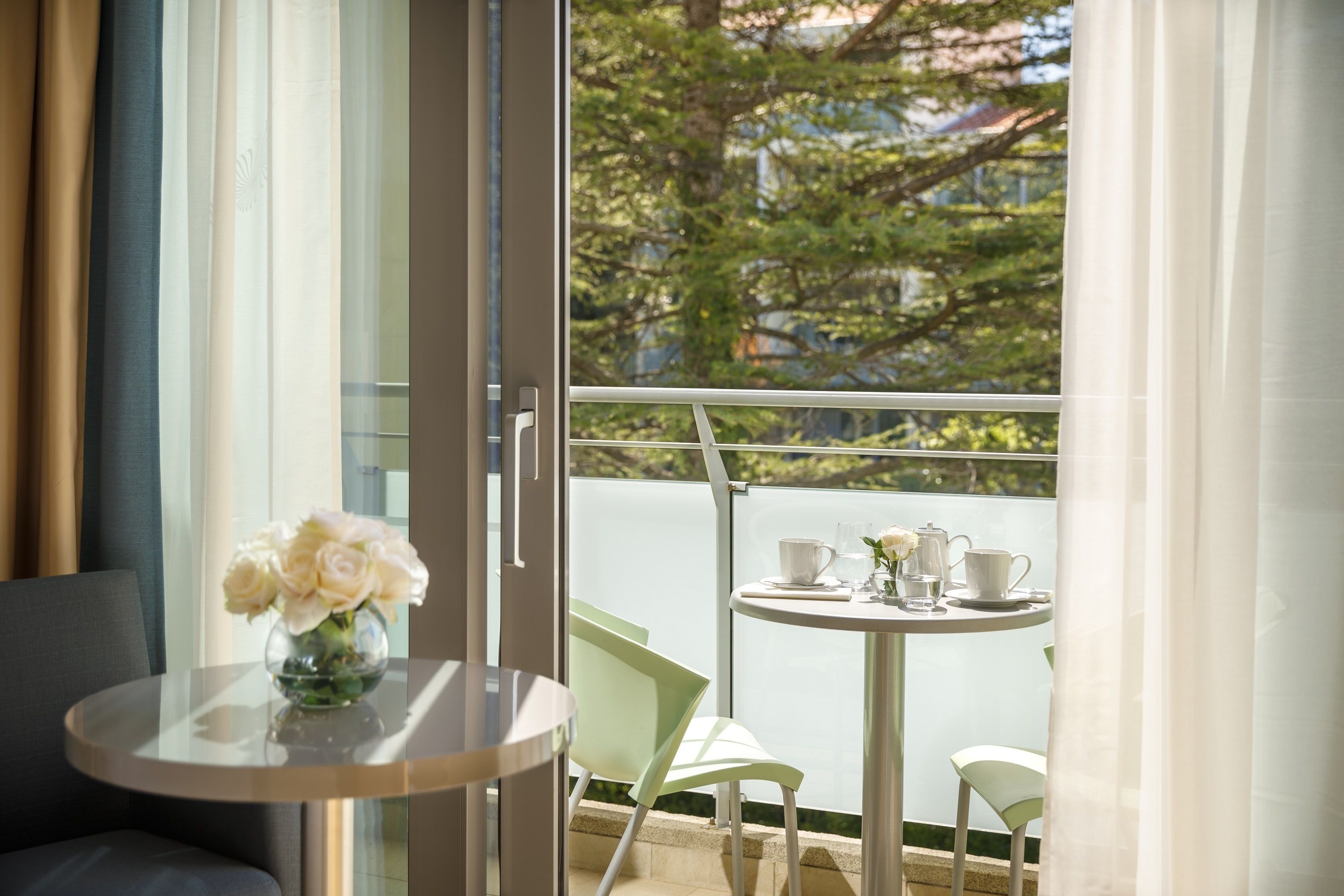 A balcony with a table set for two, a vase with flowers, and a view of trees and buildings.