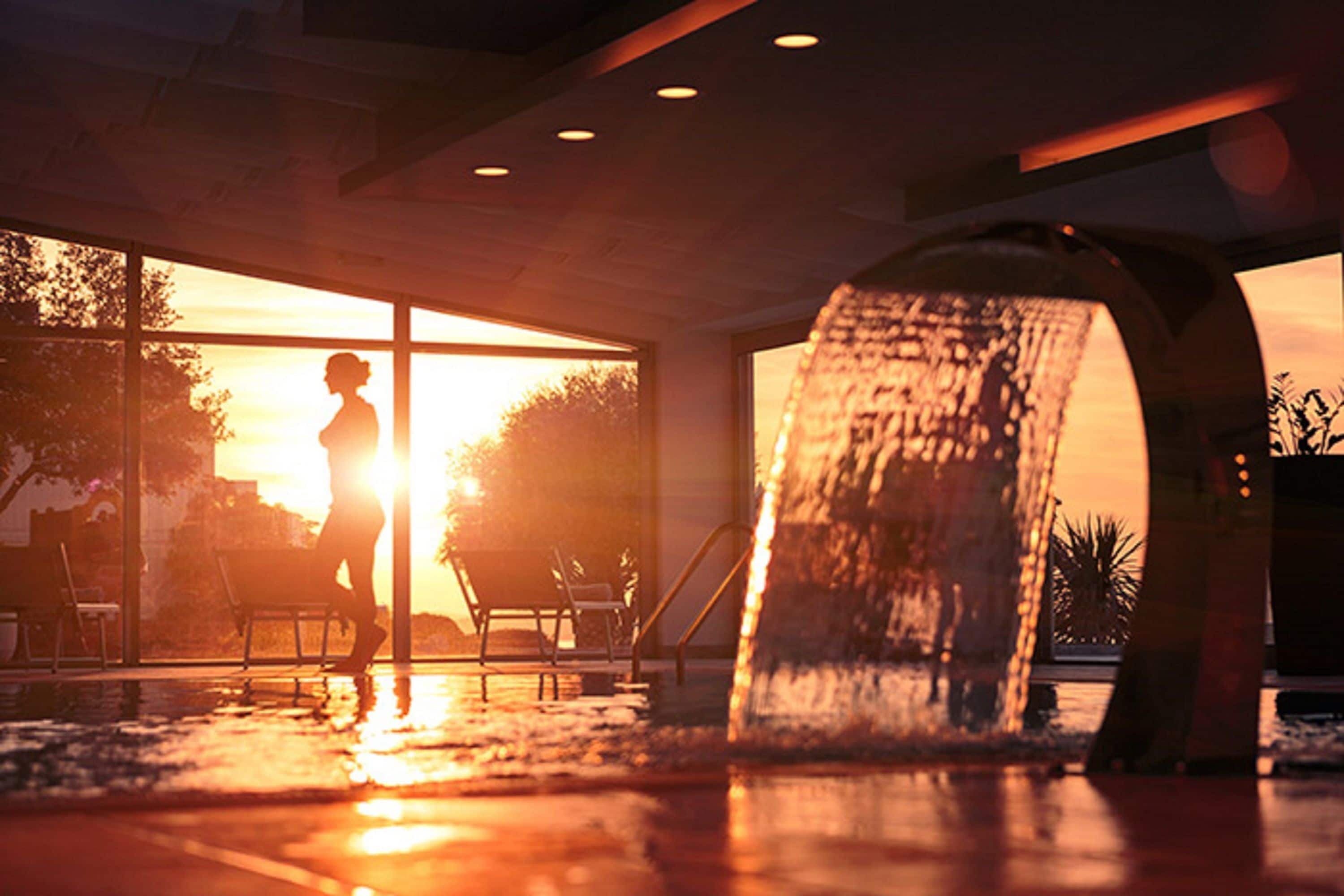 Een moderne binnenzwembadruimte met iemand die bij het zwembad staat, een waterattractie en uitzicht op de zonsondergang door grote ramen.