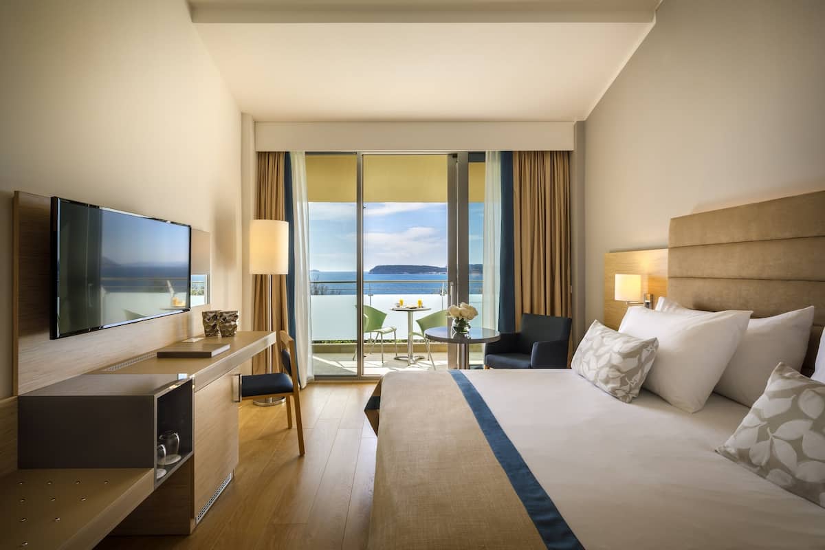 A hotel room with a bed, a desk with a flat-screen TV, a chair, a small table with a vase, and a view of the sea through large windows.