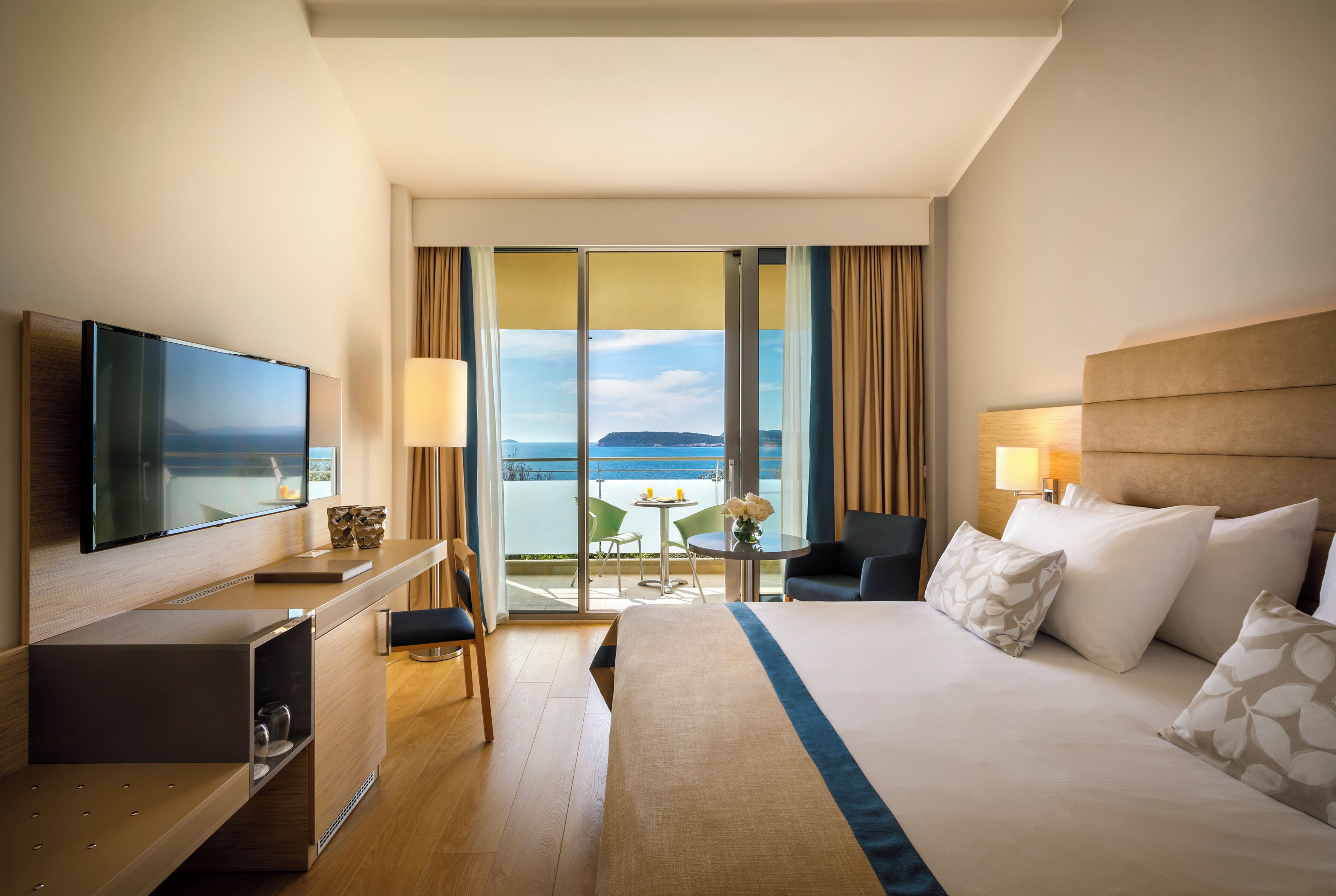 A hotel room with a bed, a desk with a TV, a balcony with a view, and a chair.