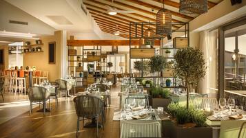 A restaurant with wooden beams, hanging wicker lamps, and a variety of plants. Tables are set with wine glasses and plates.