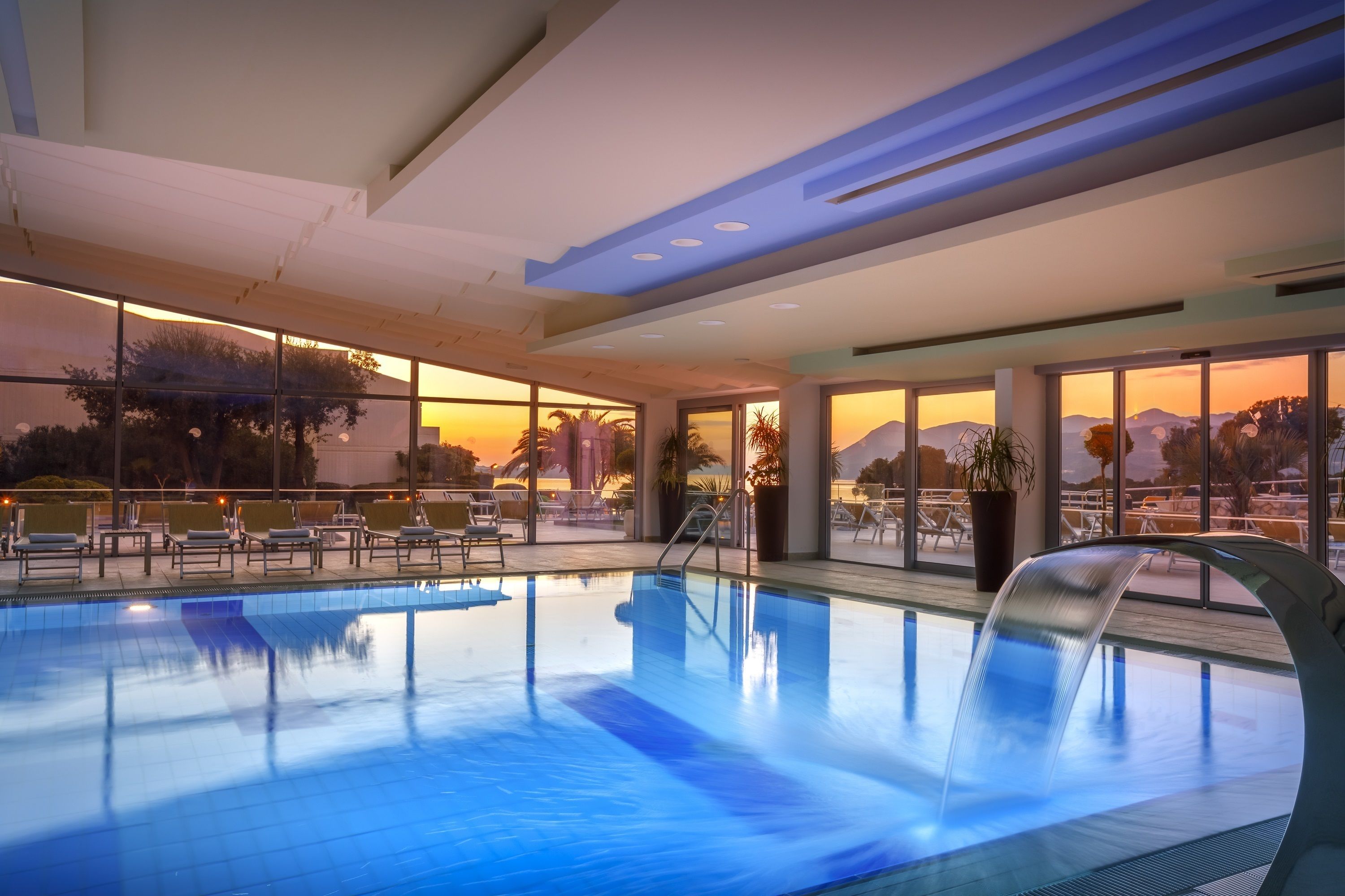 Piscina interna com paredes de vidro transparente, vista para o pôr do sol e design arquitetônico moderno.
