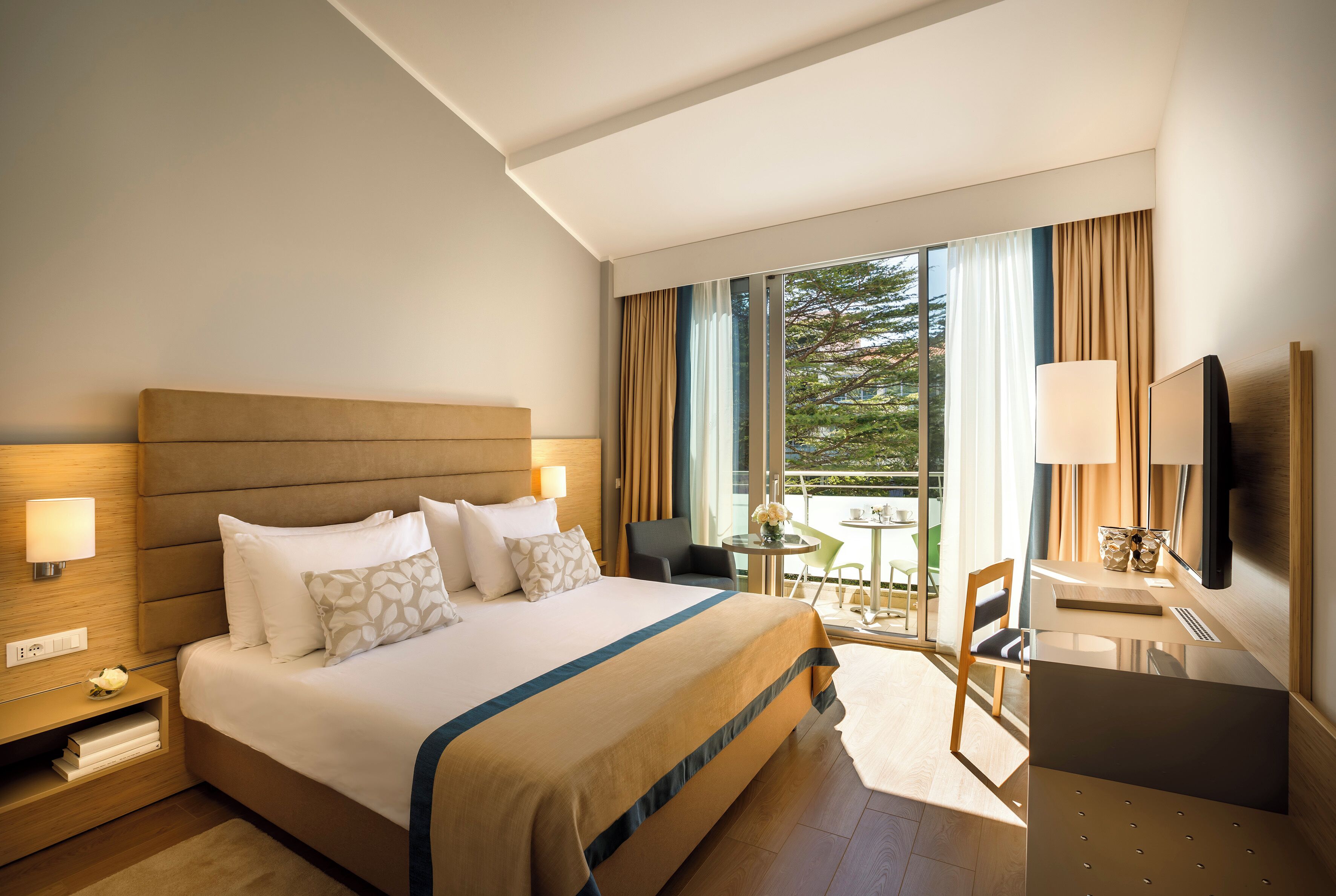 A hotel room with a large bed, bedside tables, a desk, a chair, and a TV.