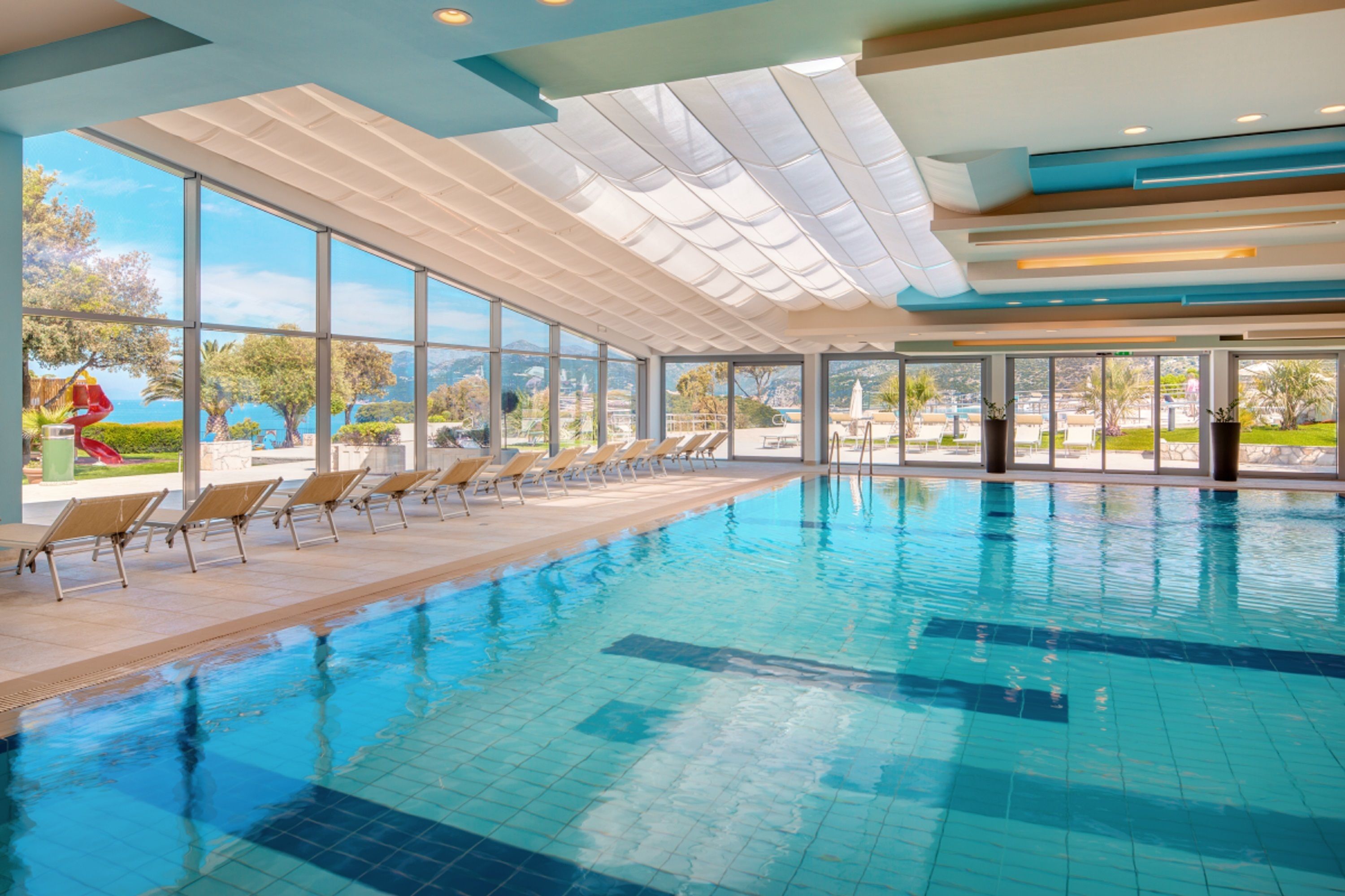 Une grande piscine intérieure aux parois en verre transparent, dotée de chaises longues et offrant une vue sur le paysage environnant.