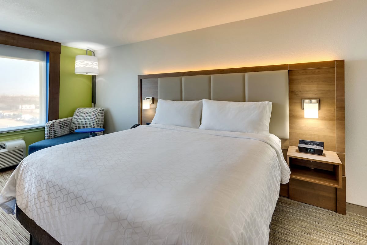 A hotel room with a large bed, a nightstand with a clock, a wall-mounted lamp, and a view of the city through the window.