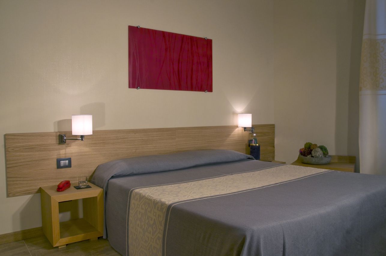 A hotel room with a double bed, two bedside lamps, a small bedside table with a lamp, a red curtain, and a wall-mounted red flag.